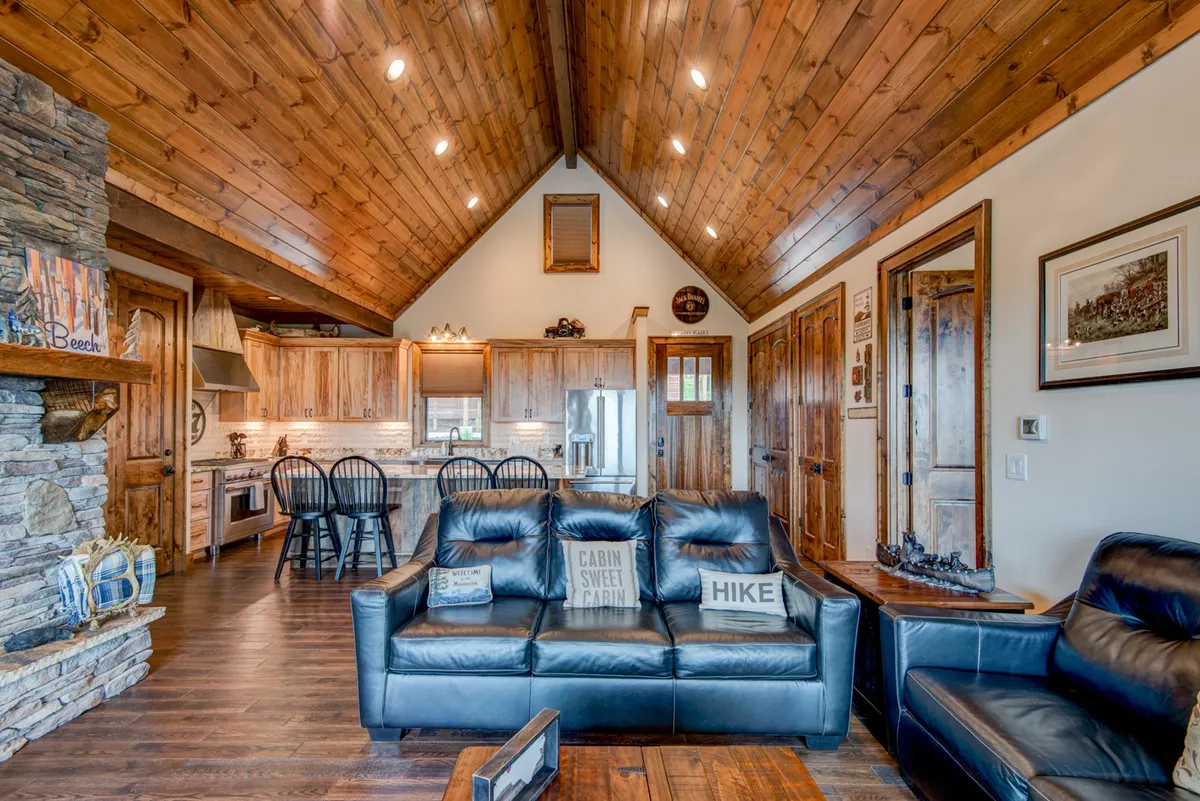 A warm, inviting interior scene with a stone fireplace, leather sofas arranged for conversation, and a view into the adjoining kitchen area. The wooden ceiling adds a rustic touch, while the overall layout facilitates relaxing, interacting, and preparing meals as a group.