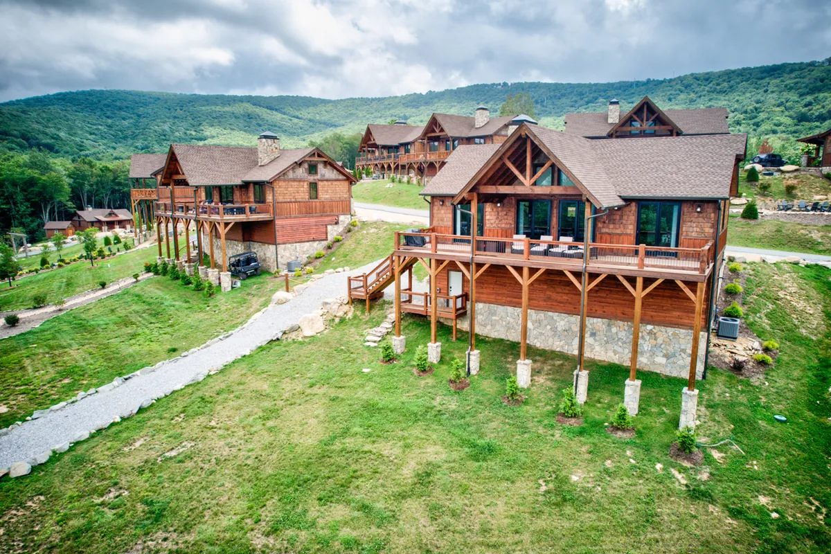 This wide exterior view showcases a cluster of wooden lodges embedded in lush greenery, framed by a stunning mountain backdrop. The surrounding landscape suggests immediate access to hiking trails and nature walks, while the large decks offer opportunities for outdoor dining and family gatherings against scenic vistas.