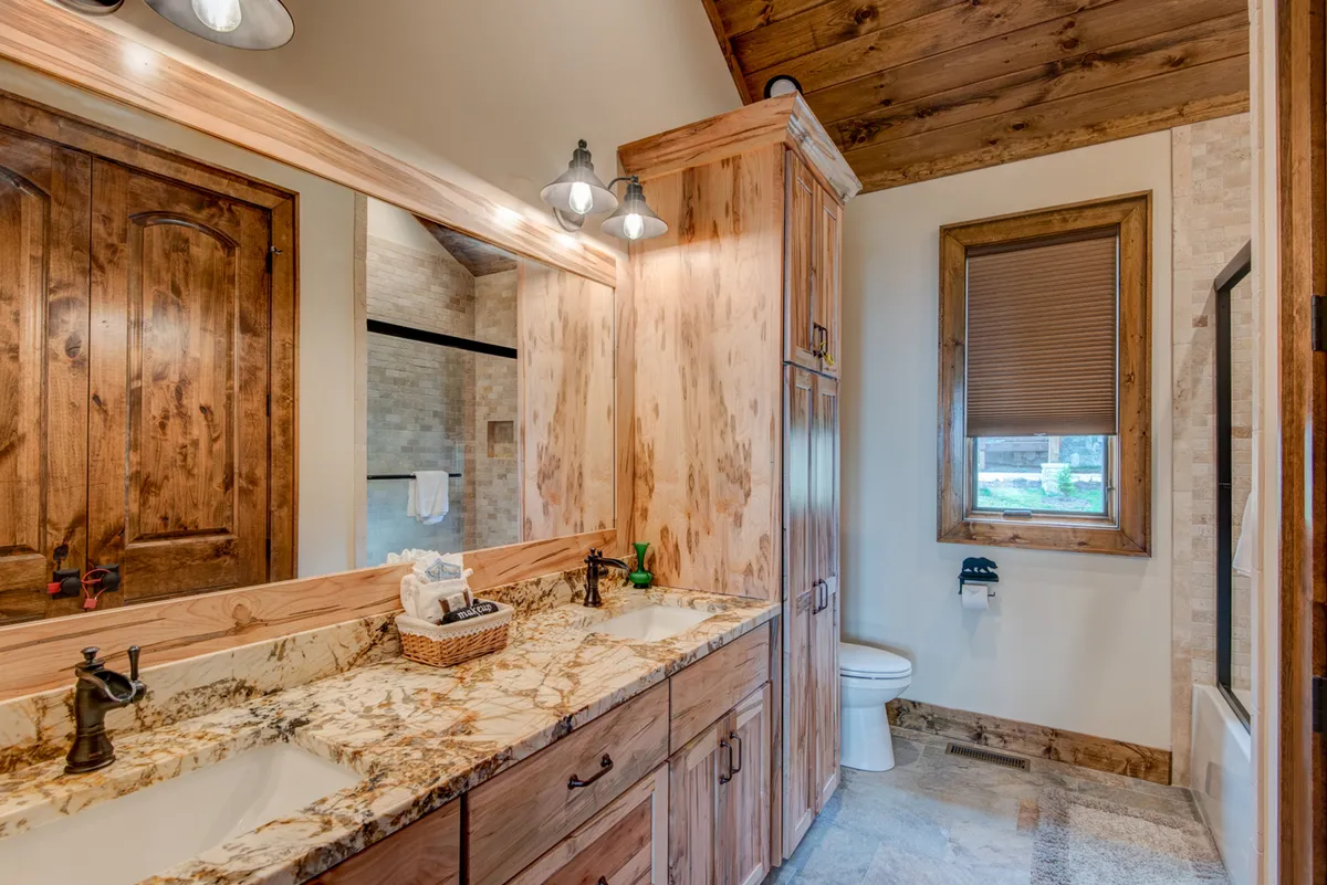 A spacious en suite bathroom featuring a double sink with granite countertop, wooden cabinetry, a large mirror, and a window framing outdoor views. The serene decor and ample natural light create a spa-like setting for personal routines.
