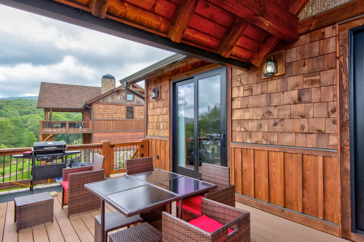 A wooden deck with an outdoor dining table and woven chairs overlooks natural scenery, suggesting a seamless blend of comfort and the outdoors. The space is set up for alfresco meals, celebrations, and casual gatherings, with a grill visible to support barbeques and group dining experiences.
