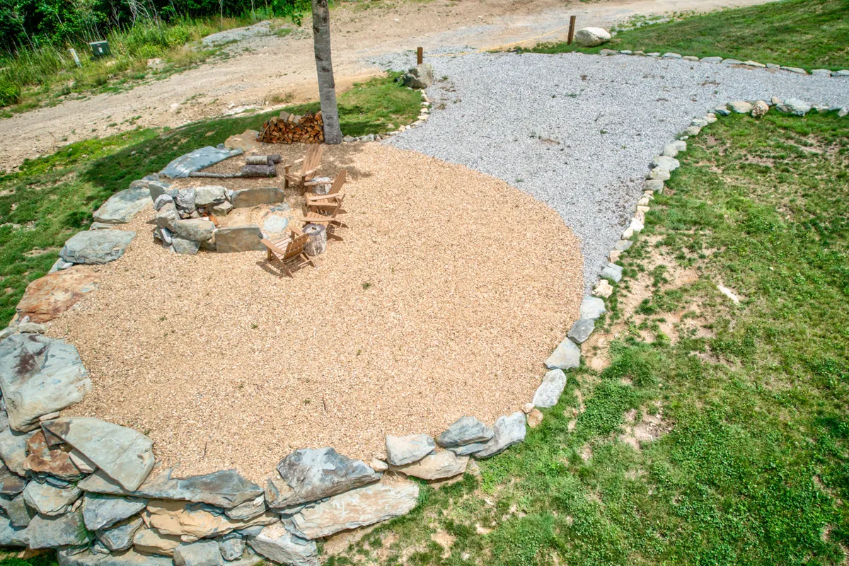 A circular stone fire pit surrounded by wooden chairs, set on a gravel pathway amidst greenery. The space is perfectly designed for gathering, making s’mores, sharing stories, and enjoying the warmth of a fire with the mountain backdrop visible beyond.