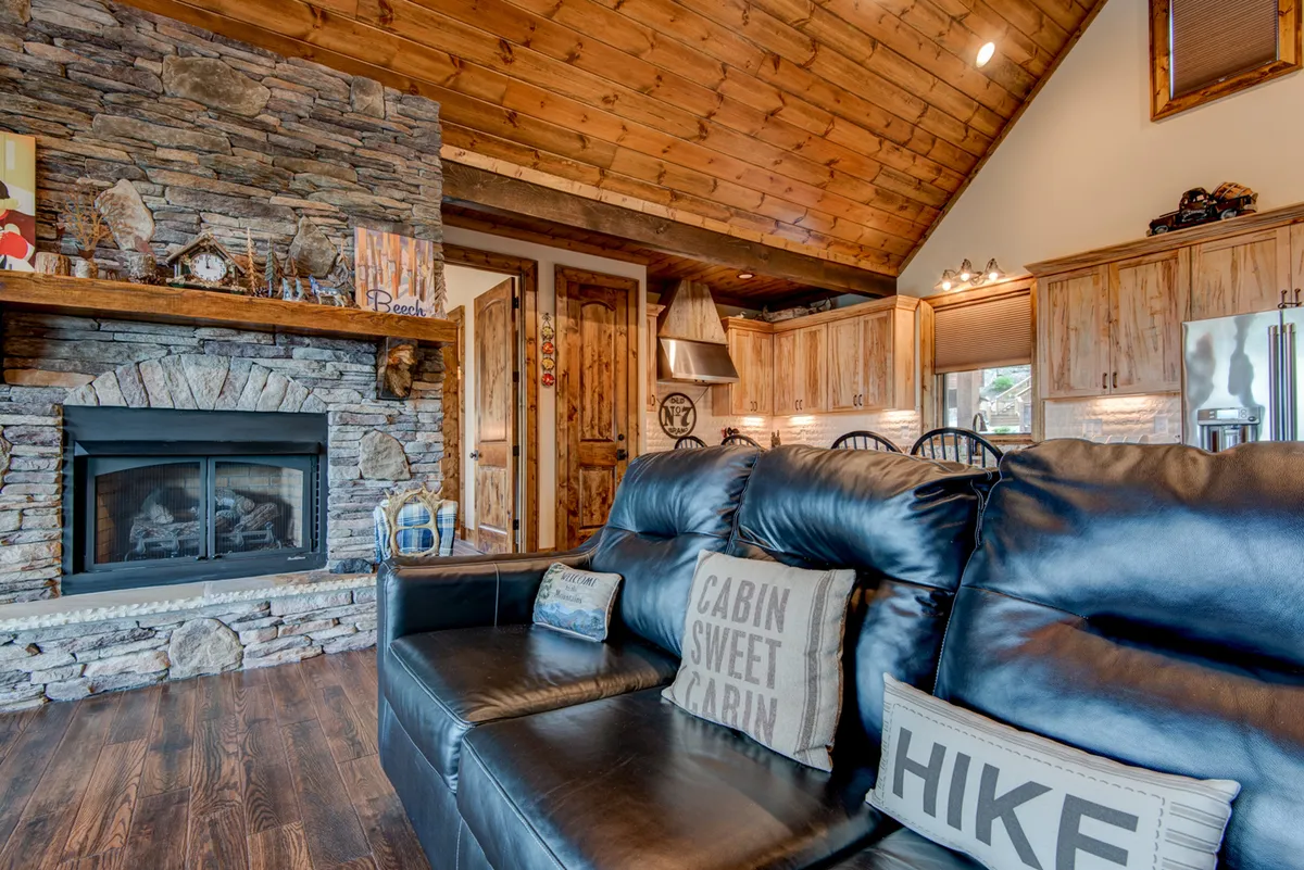 A cozy interior featuring a stone fireplace, black leather sofa, wooden cabinetry, and decorative pillows. The arrangement is ideal for both socializing and enjoying a quiet evening by the fire, with the kitchen visible in the background for a true open-concept feel.