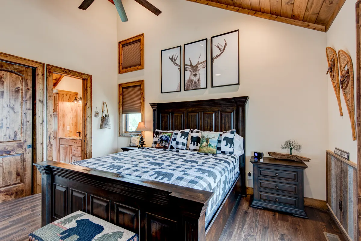 A comfortable king suite featuring a plush bed with patterned bedding, a nightstand with a lamp providing gentle lighting, framed animal artwork on the wall, and a wooden door. The space basks in soft natural light, contributing to a tranquil and rustic ambiance.