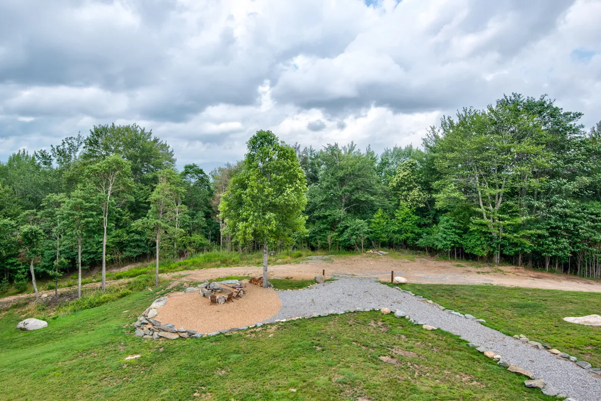 This image features a tranquil outdoor lawn with a central stone fire pit, surrounded by lush greenery and a lone tree. The gravel pathway hints at space for outdoor games and easy movement between activity areas. The open lawn is ideal for gatherings, picnics, and enjoying nature, with plenty of room for family and friends to relax after a day of adventure.