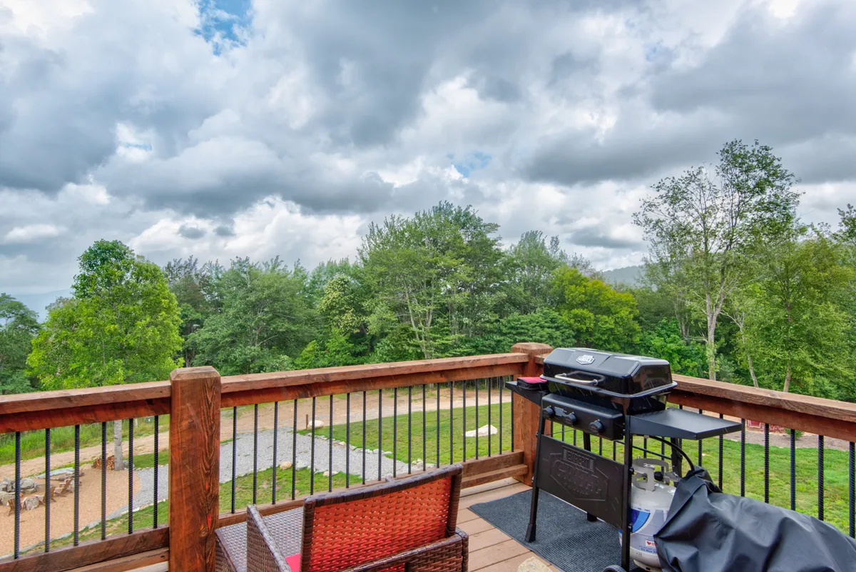 The image features a wooden railing, an outdoor grill, comfortable seating, and surrounding greenery, all positioned to make the most of the peaceful mountain environment. The scene suggests fresh air dining, relaxed afternoons, and wildlife observation in a serene setting.