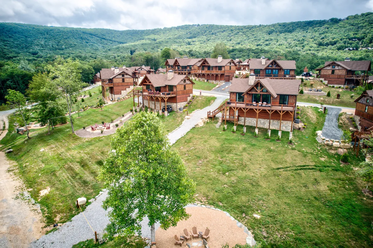 A relaxed, communal area with log cabins, a green landscape, a fire pit, and winding walking paths. Families and friends are depicted gathering for meals, enjoying campfires, and strolling through nature.
